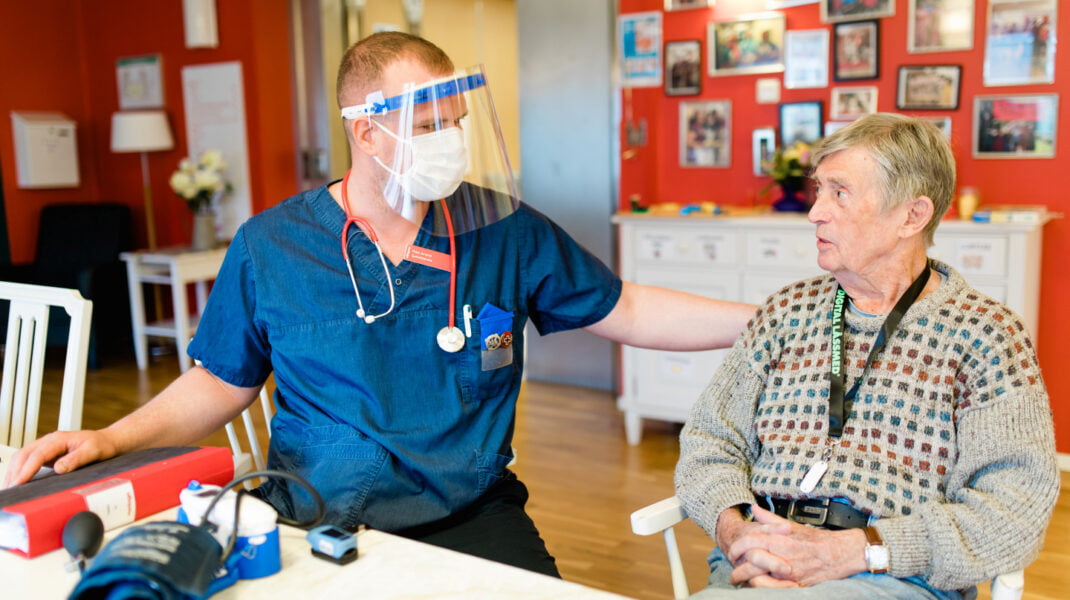Sjuksköterska med munskydd och visir samtalar med äldre man