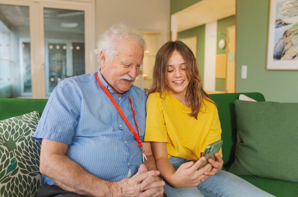 En äldre person och ett barn sitter i en soffa och tittar på en mobiltelefon