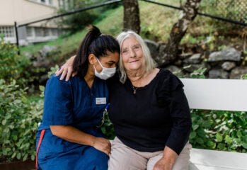 En medarbetare med munskydd omfamnar en leende omsorgstagare på en parkbänk
