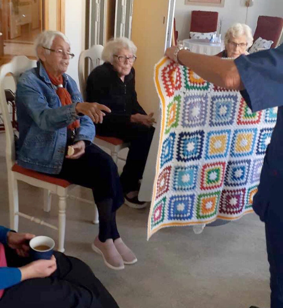 person håller upp lapptäcke framför två äldre kvinnor