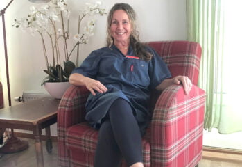 Maria i blå arbetskläder sitter i rödrutig fåtölj