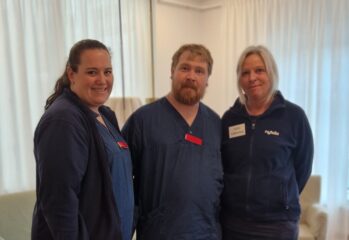 Porträtt på aktivitetspedagog Josefin, praktikant Isak och arbetshandledare Catharina. Mörkblå kläder. Står ihop, tittar in i kameran och ler.