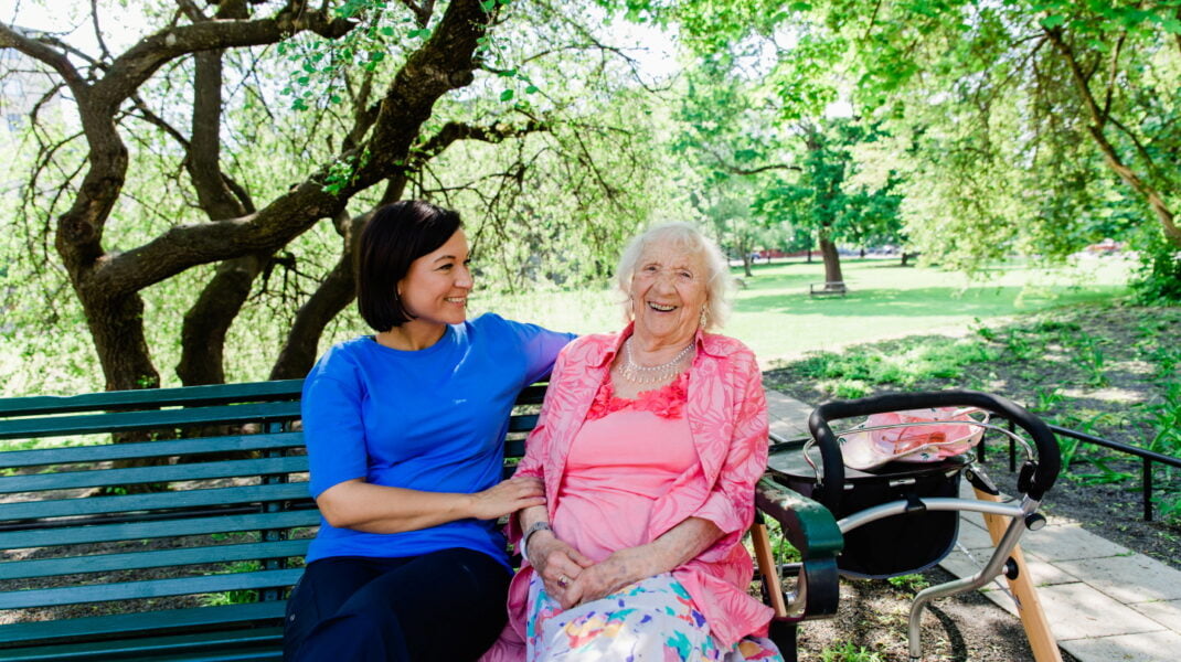Medarbetare och hemtjänstkund sitter tillsammans på en bänk utomhus och ler
