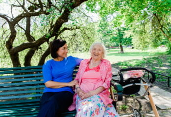 Medarbetare och hemtjänstkund sitter tillsammans på en bänk utomhus och ler
