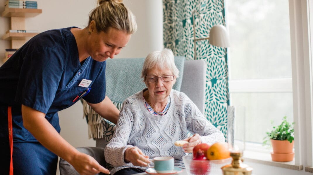 Medarbetare i blå rock lutar sig över boende som sitter i fåtölj vid bord med kaffekopp