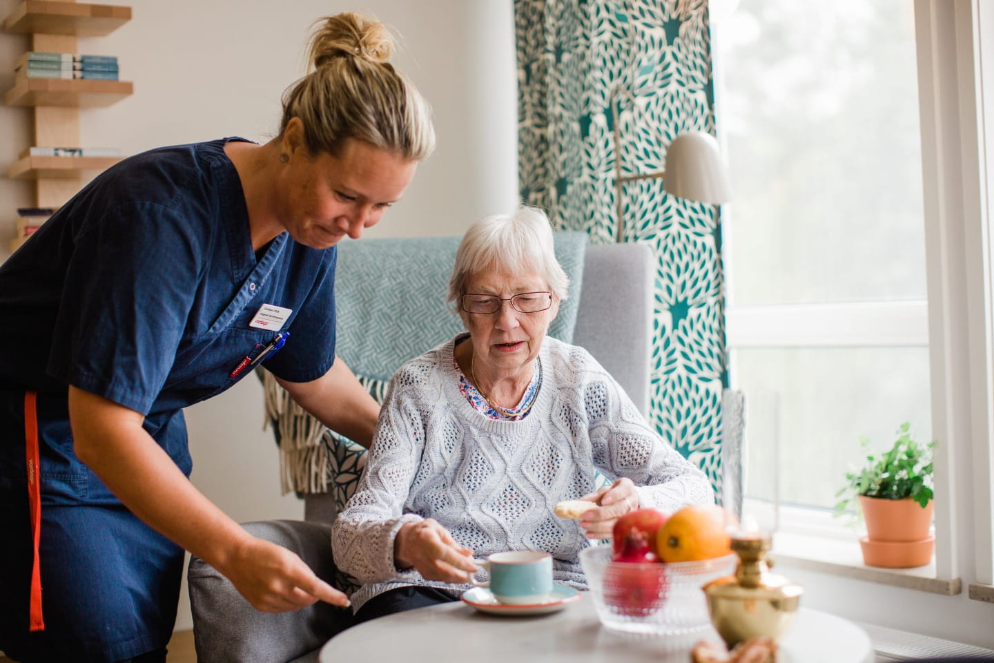 Medarbetare i blå rock lutar sig över boende som sitter i fåtölj vid bord med kaffekopp
