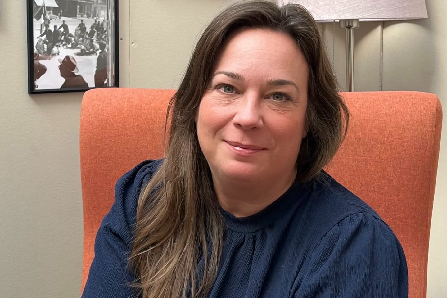 Porträtt av en kvinna som sitter i en orange fåtölj och ler mot kameran, i en hemtrevlig miljö med tavlor och lampa i bakgrunden.