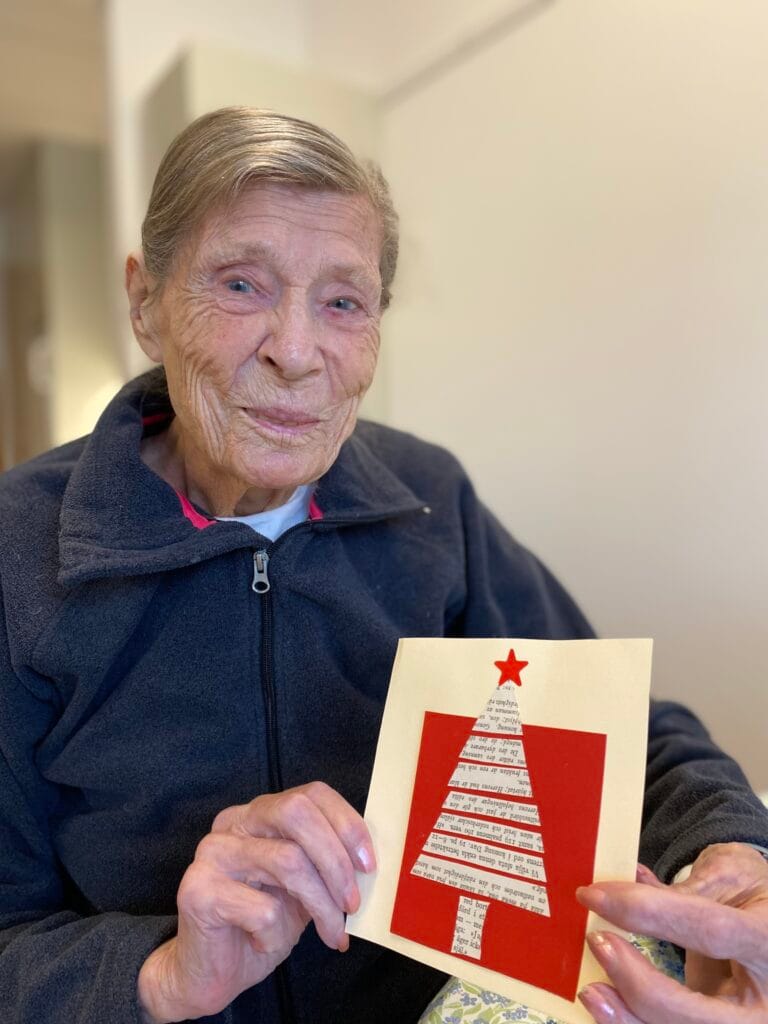 En äldre kvinna sitter inomhus och håller upp ett handgjort julkort med en gran av tidningspapper på röd bakgrund och en röd stjärna högst upp. Hon ler varmt mot kameran och bär en mörk fleecejacka.