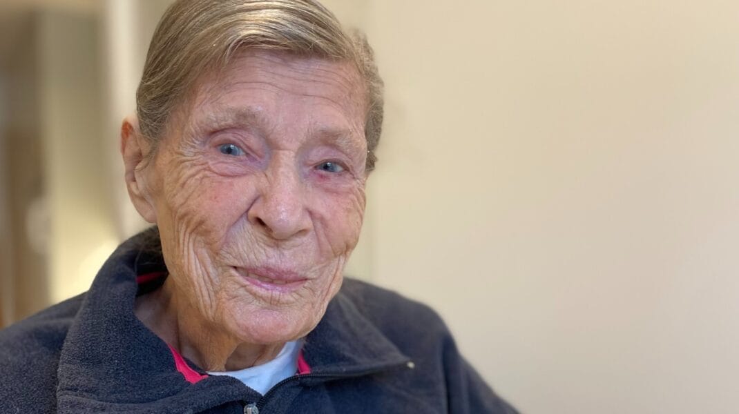 En äldre kvinna sitter inomhus och håller upp ett handgjort julkort med en gran av tidningspapper på röd bakgrund och en röd stjärna högst upp. Hon ler varmt mot kameran och bär en mörk fleecejacka.