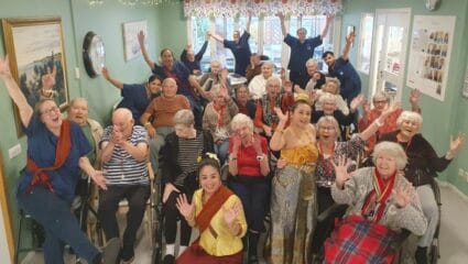 En stor grupp boende och vårdpersonal sitter tillsammans i ett ljust rum och ler och vinkar mot kameran. Ljusslingor och tavlor pryder väggarna i bakgrunden.