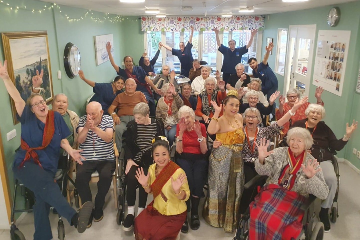 En stor grupp boende och vårdpersonal sitter tillsammans i ett ljust rum och ler och vinkar mot kameran. Ljusslingor och tavlor pryder väggarna i bakgrunden.