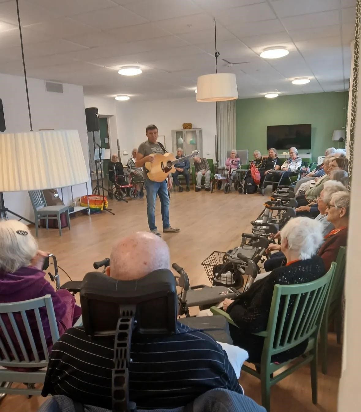 En man står med en gitarr och uppträder inför en grupp boende i ett gemensamt rum. Publiken sitter i stolar och rullstolar och lyssnar.