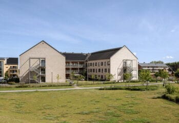 Exteriörbild av äldreboendet Villa Vikhem i Staffanstorp, en modern ljus tegelbyggnad omgiven av grönskande parkmiljö och gångvägar under en klarblå himmel.
