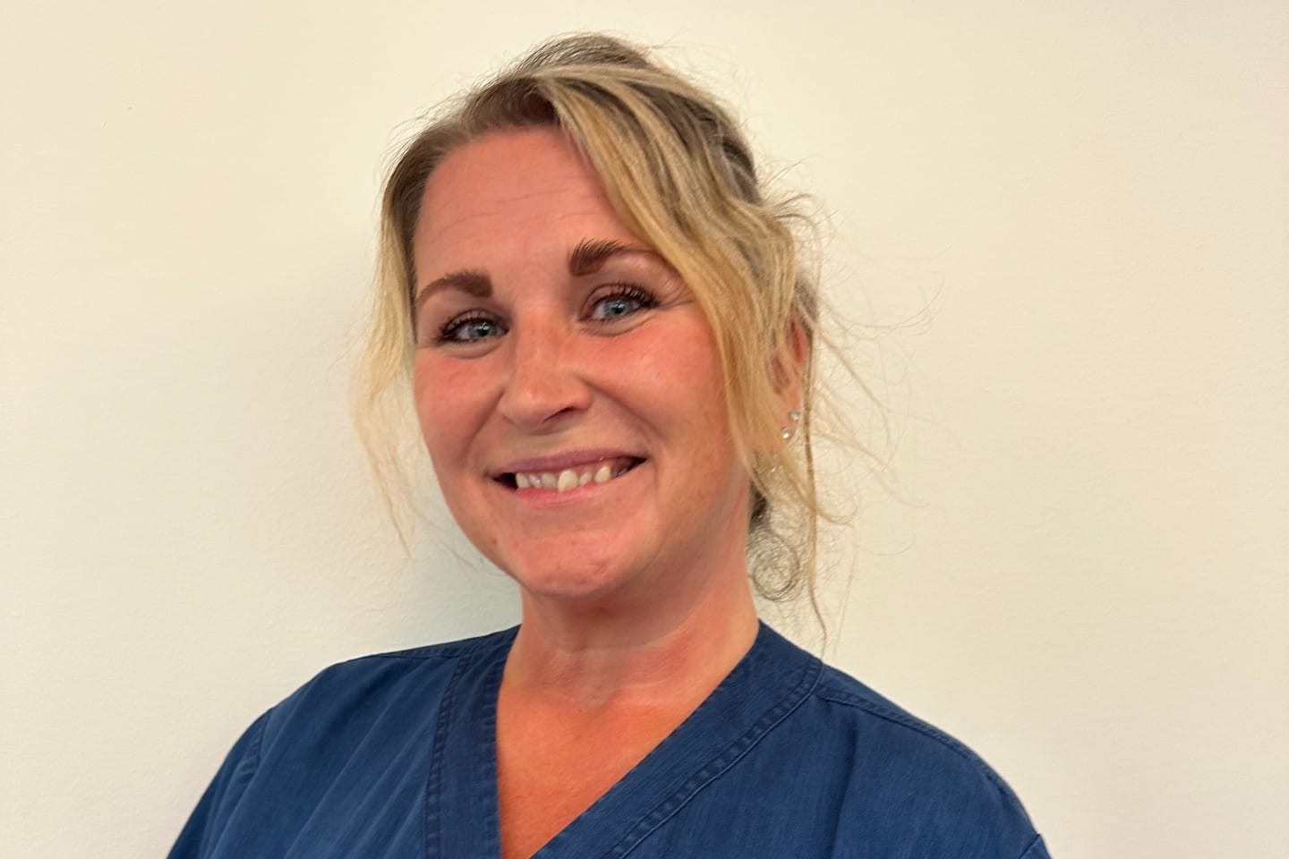 Porträtt av en leende kvinna i blå vårdklädsel framför en ljus vägg, fotograferad i närbild inomhus