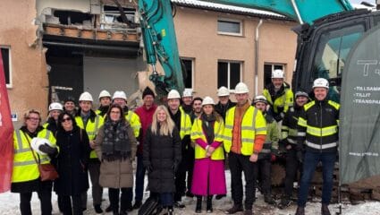 En större grupp personer i varselkläder och hjälmar står framför en byggnad som delvis rivits, med en grävmaskin bakom sig och snö på marken.