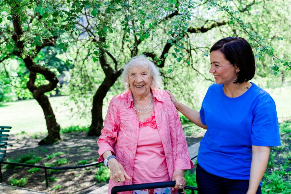 Äldre kvinna med rollator promenerar i en park tillsammans med en medarbetare.