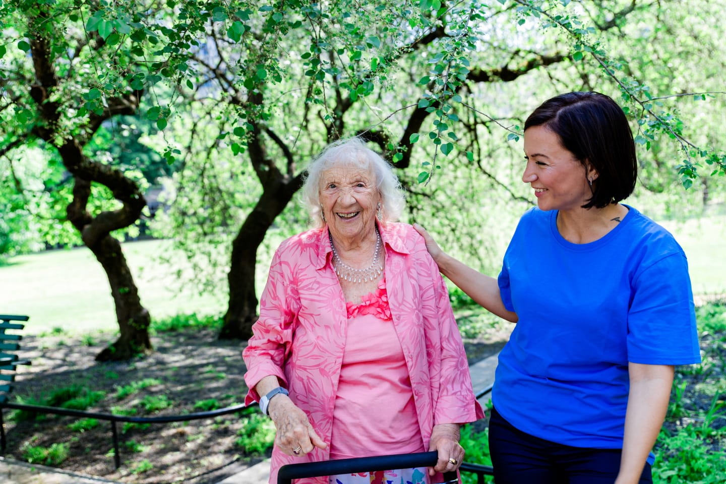 Äldre kvinna med rollator promenerar i en park tillsammans med en medarbetare.