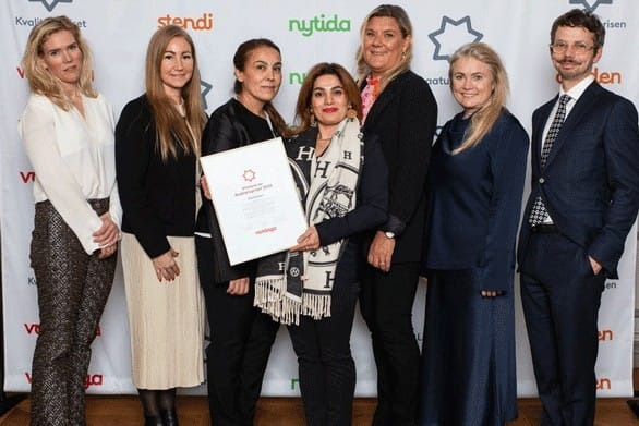 n grupp på sju personer står tillsammans framför en bakgrund med logotyper och poserar för bild. En person i mitten håller upp ett diplom från Kvalitetspriset, och alla är klädda i formella eller semi-formella kläder.