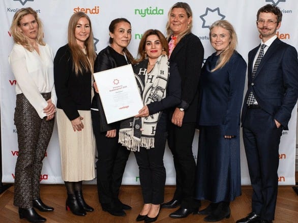 En grupp på sju personer står tillsammans framför en bakgrund med logotyper och poserar för bild. En person i mitten håller upp ett diplom från Kvalitetspriset, och alla är klädda i formella eller semi-formella kläder.