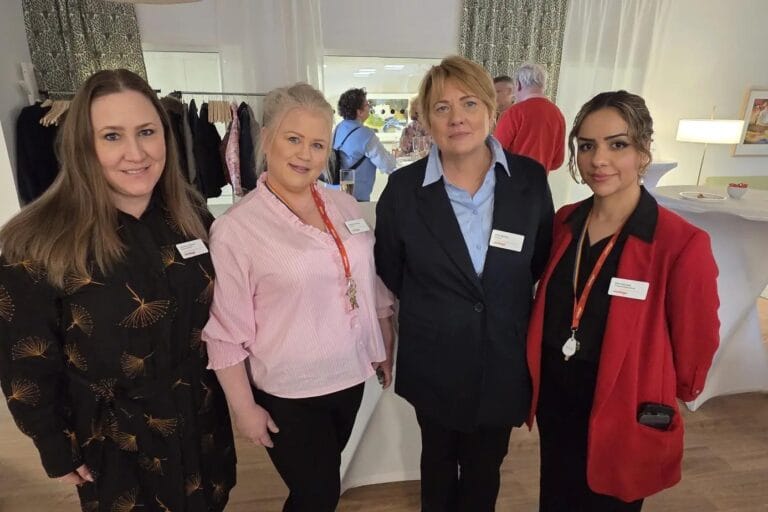 Fyra kvinnor står sida vid sida och ler mot kameran i en ljus och hemtrevlig inomhusmiljö. De bär namnskyltar och står framför ett bord med kläder och andra personer i bakgrunden, vilket ger intryck av ett evenemang eller en träff i gemensamhetslokal.