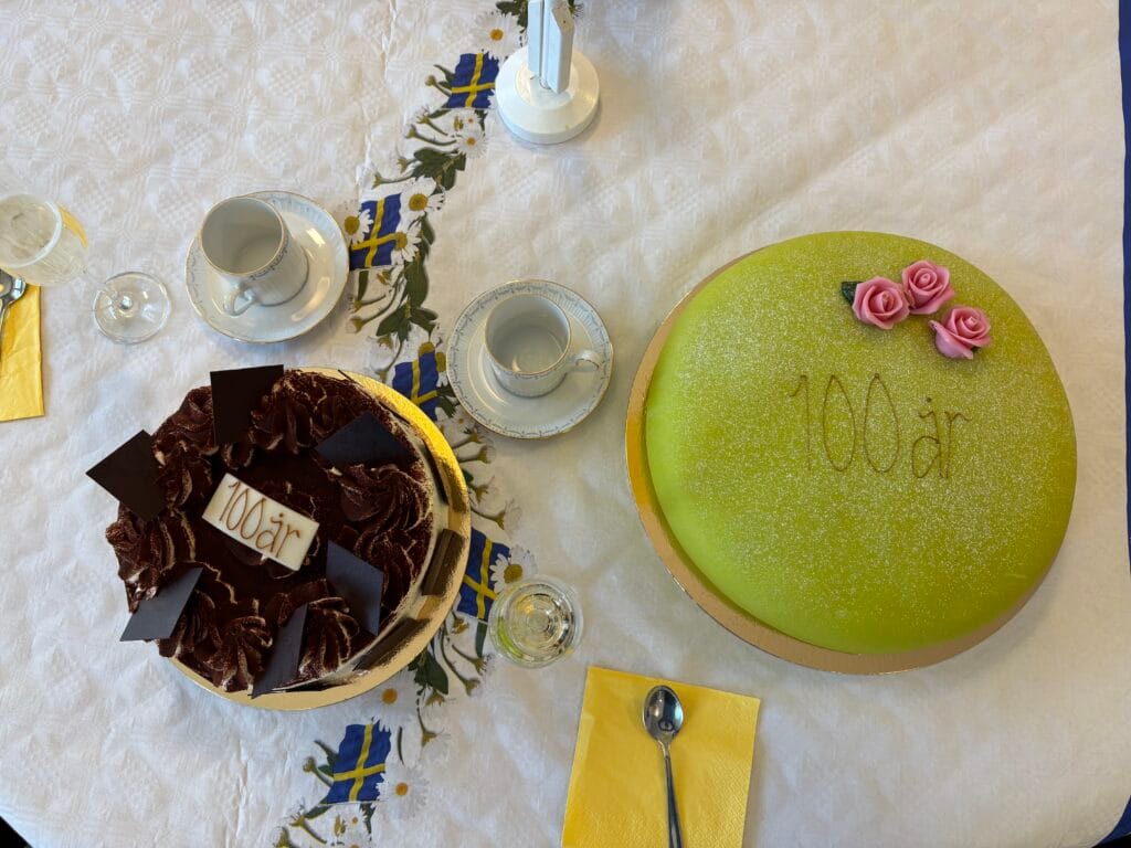 Två tårtor på dukat bord, dekorerade med texten “100 år” samt svenska flaggor och kaffekoppar runtom.