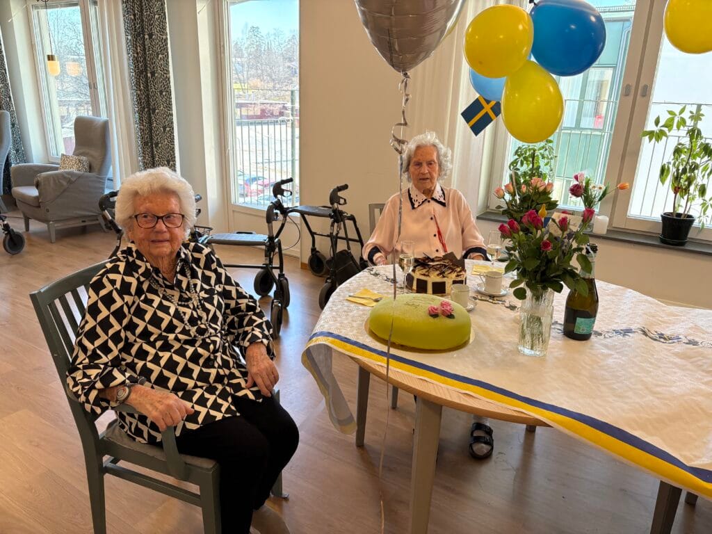 Två äldre kvinnor sitter vid ett festdukad bord med tårtor, blommor och blågula ballonger i ett ljust rum.