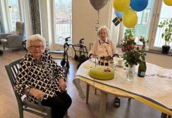Två äldre kvinnor sitter vid ett festdukad bord med tårtor, blommor och blågula ballonger i ett ljust rum.