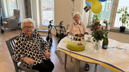 Två äldre kvinnor sitter vid ett festdukad bord med tårtor, blommor och blågula ballonger i ett ljust rum.