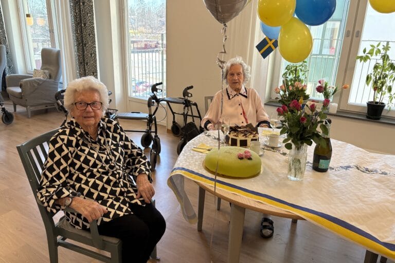 Två äldre kvinnor sitter vid ett festdukad bord med tårtor, blommor och blågula ballonger i ett ljust rum.