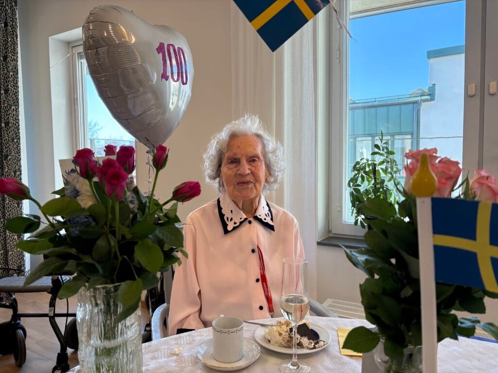 Äldre kvinna firar 100 år och sitter vid bord med tårta, blommor och en ballong formad som ett hjärta med texten “100”.