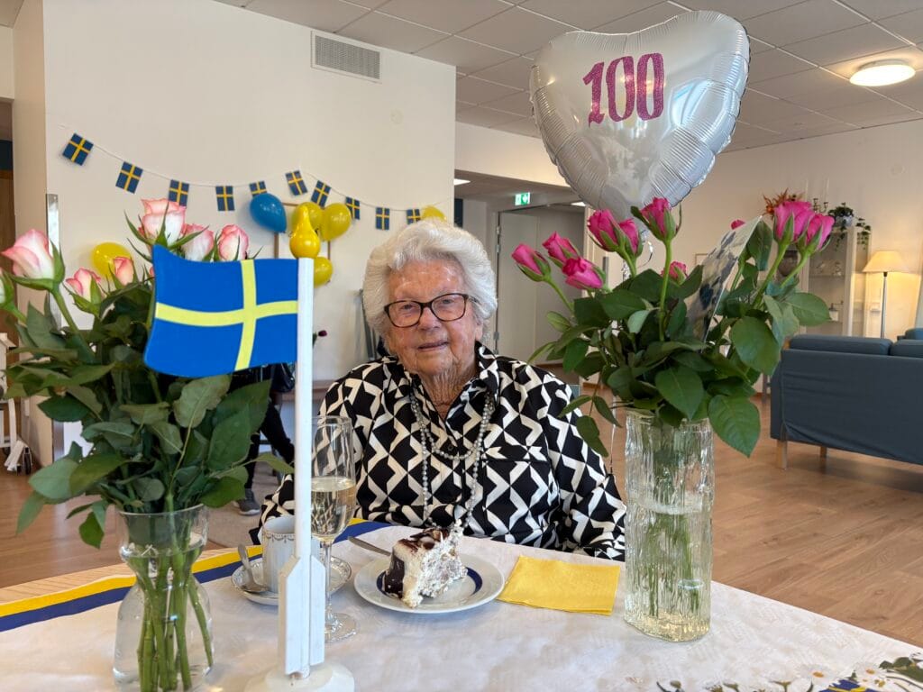 Äldre kvinna sitter vid festdukad plats med tårta och blommor, omgiven av svenska flaggor och en ballong med texten “100”.