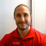 Närbild på en leende man i röd pikétröja med namnbricka, stående inomhus mot en ljus bakgrund.