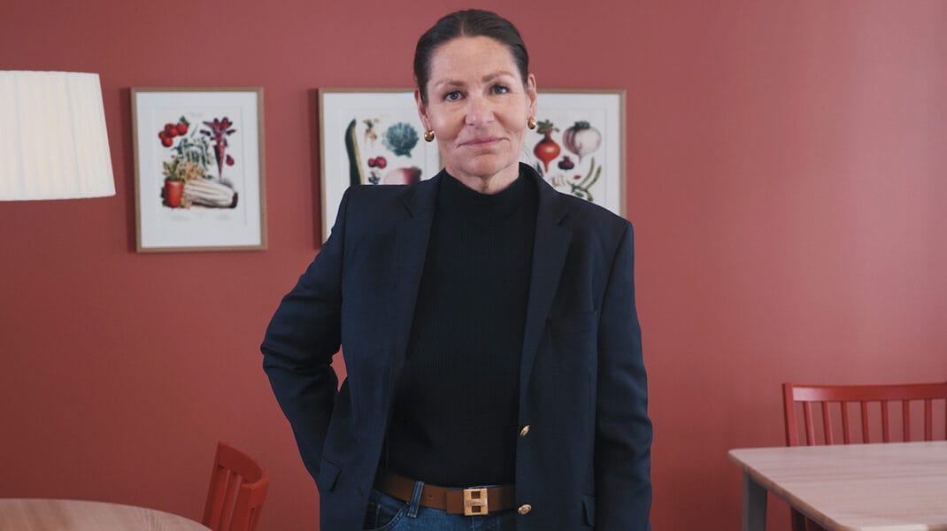 Kvinna i mörk kavaj och svart tröja står i ett ljust rum med röd vägg, träbord och stolar, samt två inramade illustrationer på väggen bakom.