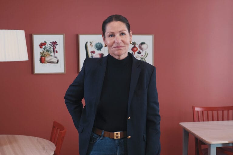 Kvinna i mörk kavaj och svart tröja står i ett ljust rum med röd vägg, träbord och stolar, samt två inramade illustrationer på väggen bakom.