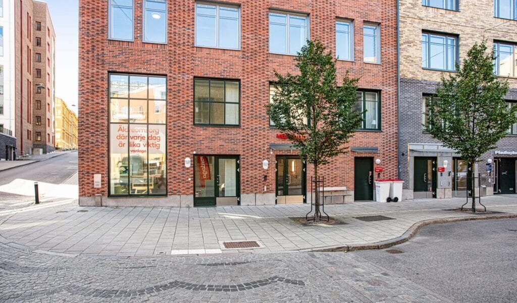Exteriör av modern tegelbyggnad i stadsmiljö med entréer i markplan, stora fönster, träd längs trottoaren och kullerstensgata i förgrunden.