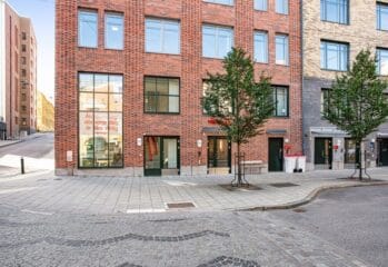 Exteriör av modern tegelbyggnad i stadsmiljö med entréer i markplan, stora fönster, träd längs trottoaren och kullerstensgata i förgrunden.