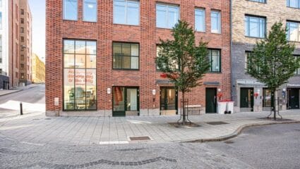 Exteriör av modern tegelbyggnad i stadsmiljö med entréer i markplan, stora fönster, träd längs trottoaren och kullerstensgata i förgrunden.