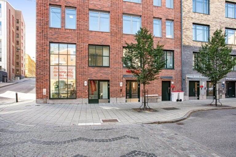 Exteriör av modern tegelbyggnad i stadsmiljö med entréer i markplan, stora fönster, träd längs trottoaren och kullerstensgata i förgrunden.