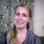 Porträtt av en leende kvinna med ljust hår, sittande inomhus framför ett mönstrat tyg i gröna toner.