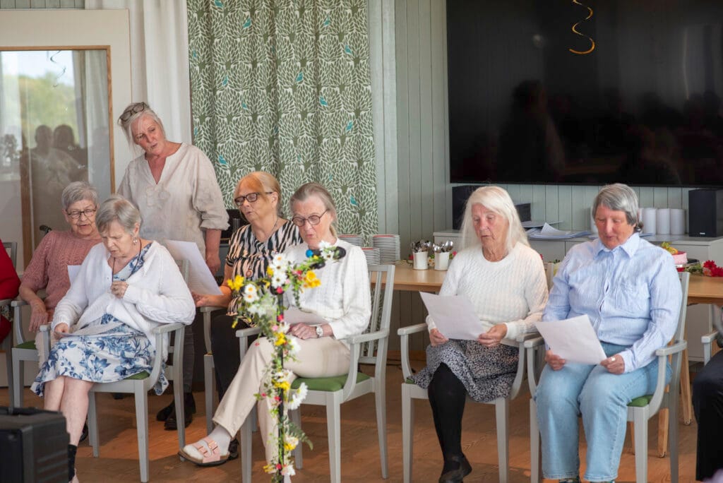 Äldre personer sitter tillsammans i ett rum och sjunger från papper