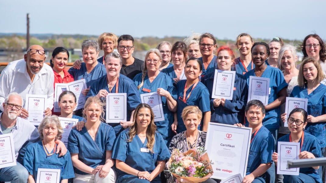 Stor grupp medarbarbetare i vårdkläder står utomhus för gruppbild med diplom och certificat