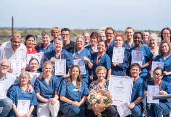 Stor grupp medarbarbetare i vårdkläder står utomhus för gruppbild med diplom och certificat