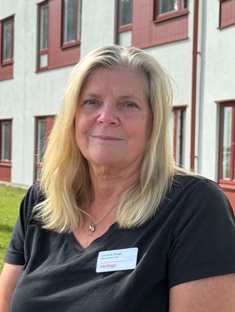 Porträttfoto av en person med axellångt ljust hår som sitter utomhus framför en byggnad med ljus fasad och röda fönsterdetaljer. Personen bär en mörk kortärmad tröja och en namnbricka. I bakgrunden syns en gräsmatta och delar av byggnaden.