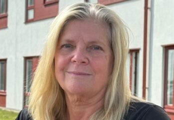 Porträttfoto av en person med axellångt ljust hår som sitter utomhus framför en byggnad med ljus fasad och röda fönsterdetaljer. Personen bär en mörk kortärmad tröja och en namnbricka. I bakgrunden syns en gräsmatta och delar av byggnaden.