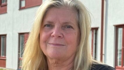 Porträttfoto av en person med axellångt ljust hår som sitter utomhus framför en byggnad med ljus fasad och röda fönsterdetaljer. Personen bär en mörk kortärmad tröja och en namnbricka. I bakgrunden syns en gräsmatta och delar av byggnaden.