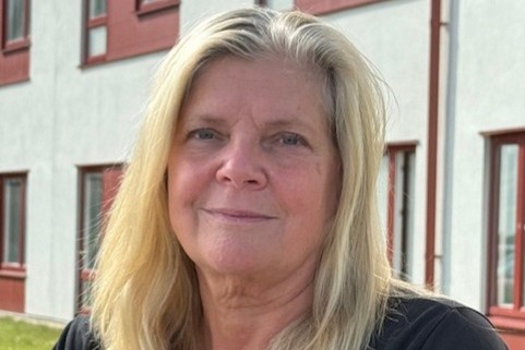 Porträttfoto av en person med axellångt ljust hår som sitter utomhus framför en byggnad med ljus fasad och röda fönsterdetaljer. Personen bär en mörk kortärmad tröja och en namnbricka. I bakgrunden syns en gräsmatta och delar av byggnaden.