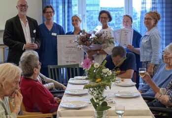 Personer samlas kring ett dukat bord i en gemensamhetslokal för att uppmärksamma en utmärkelse. Diplom och blommor hålls upp medan boende sitter runt bordet.