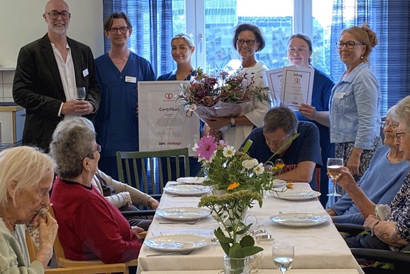 Personer samlas kring ett dukat bord i en gemensamhetslokal för att uppmärksamma en utmärkelse. Diplom och blommor hålls upp medan boende sitter runt bordet.
