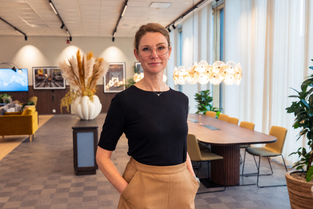 Porträtt av en person med glasögon och mörk tröja som står i en modern inomhusmiljö med stora fönster, växter och dekorativ belysning i bakgrunden.