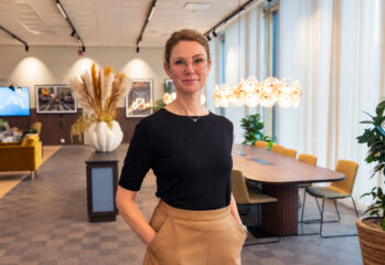 Porträtt av en person med glasögon och mörk tröja som står i en modern inomhusmiljö med stora fönster, växter och dekorativ belysning i bakgrunden.